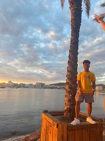 Male student stands next to a body of water and a palm tree in Spain