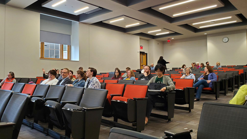 Audience seated in a lecture hall with red and black chairs under bright ceiling lights.