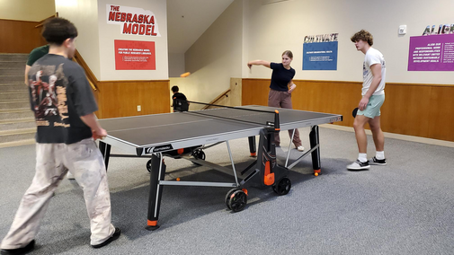 Four people playing ping pong in a brightly lit indoor space with posters on the walls.