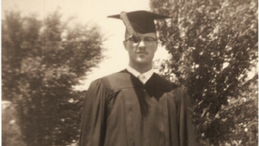 Man in graduation cap and gown outdoors, trees in the background.