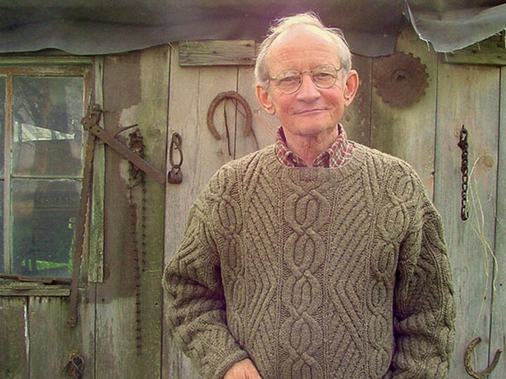 Ted Kooser a knit sweater holding a cup, standing in front of an old wooden shed with rustic tools.