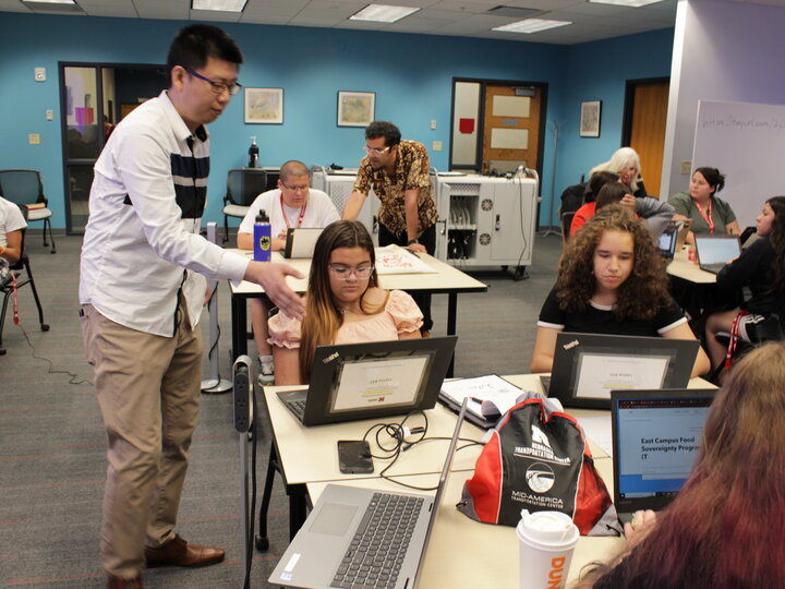 Librarians assisting students with laptops in a classroom around tables of 4.
