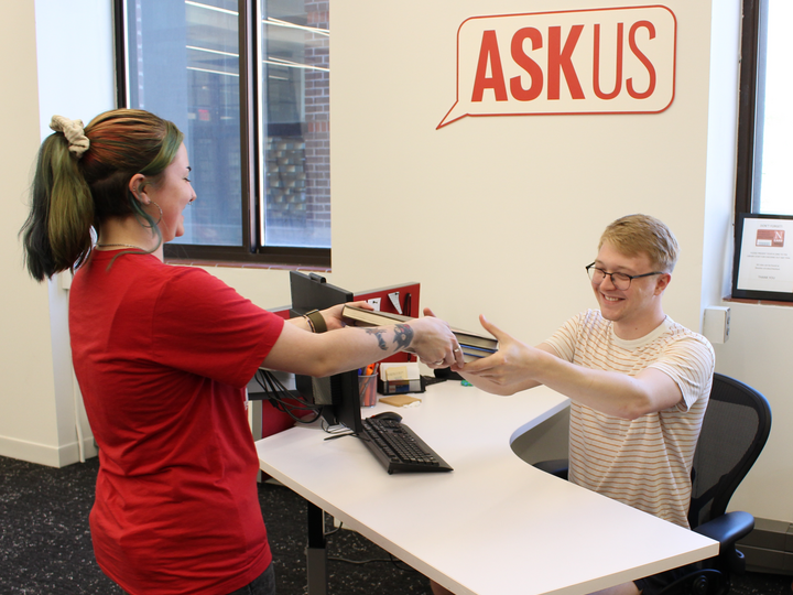 Student handing books to an ASK us person sitting behind a desk