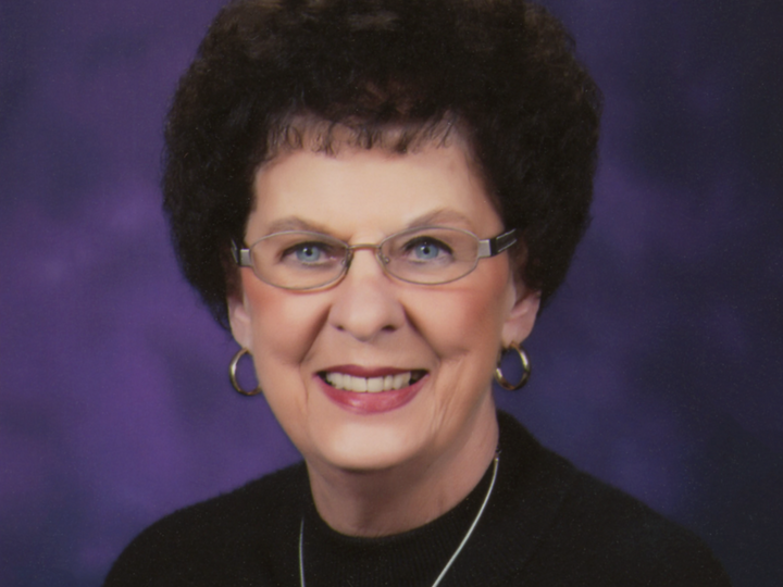 Helen Plasters Nemzek with glasses, curly dark hair, wearing a black top against a purple background.