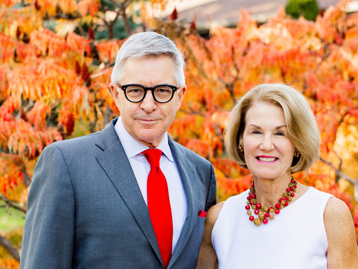 Man in a gray suit and woman in a white dress smiling in front of autumn foliage.