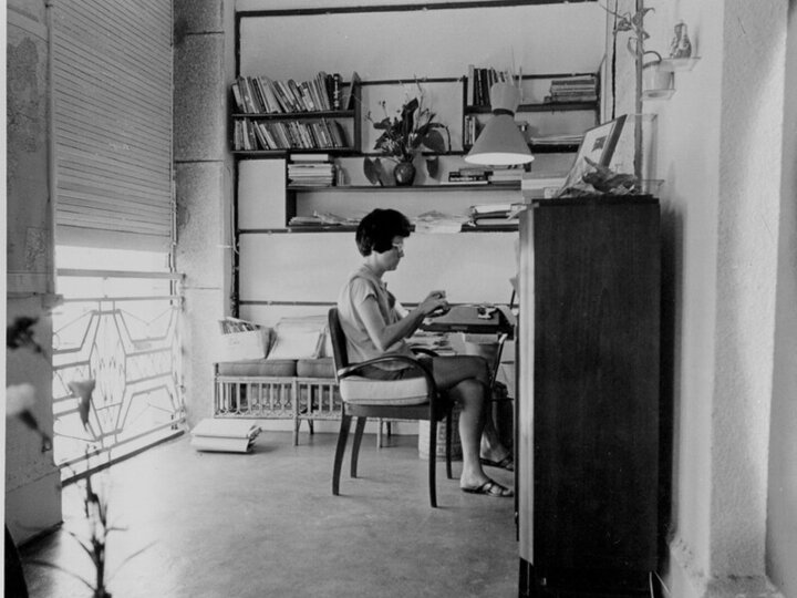Beverly Keever in her apartment sitting at a desk with a window behind her typing into a typewriter