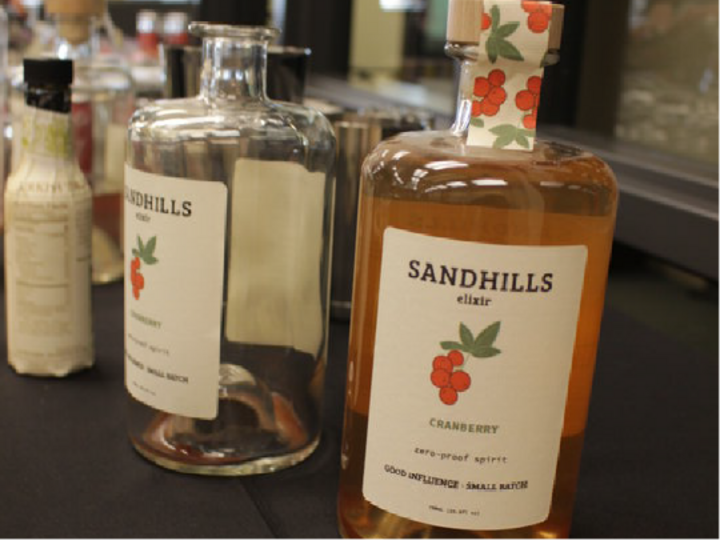 Sandhills cranberry elixir bottles on a table with other beverages in the background.