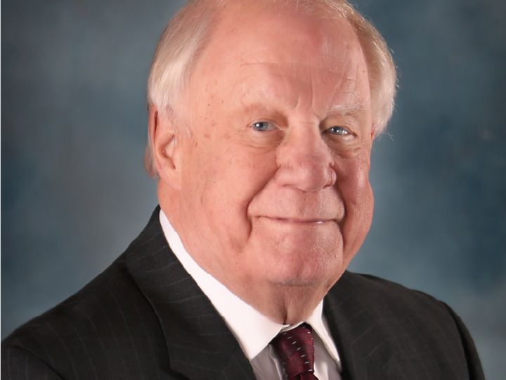 Stuart Embury in suit and tie with a neutral expression, set against a blue gradient background.