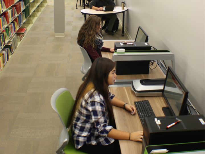 People studying at desks in a library with bookshelves along one side.