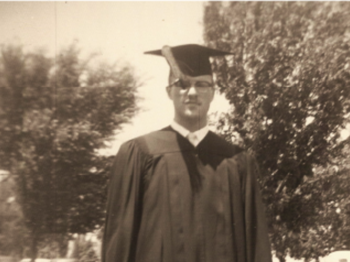 Man in graduation cap and gown outdoors, trees in the background.