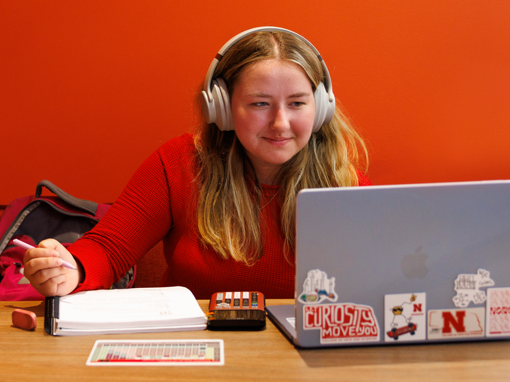 Student wearing headphones, studying at a laptop with stickers, notes, and a calculator on the table.