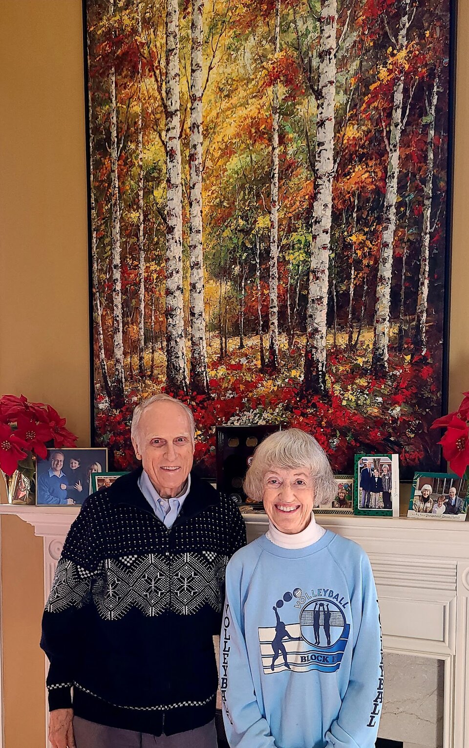 Ray and Roby Schmidt smiling in front of a mantel with photos and a painting of autumn birch trees.